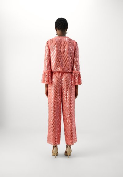 Pink sequin jumpsuit with long sleeves and ruffled cuffs, featuring a cinched waist and wide-leg pants. Worn with nude high heels.