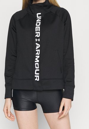 Femme portant une veste zippée noire Under Armour avec un logo vertical et un short de sport noir ajusté, debout devant un fond uni.