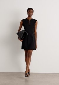 Black sleeveless dress with a keyhole neckline and side pocket. Model holds a black clutch, wearing flat sandals on a neutral background.