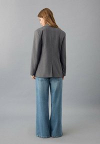 Grijze blazer met krijtstreep en gestructureerde schouders, gecombineerd met lichtblauwe wijde broekspijpen met hoge taille. Soepele stof en een nauwsluitende pasvorm.