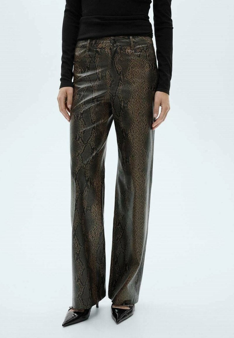 Femme portant un pantalon taille haute imprimé cuir de serpent marron et noir brillant, associé à des talons noirs pointus et un haut noir cintré à manches longues.