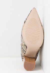 Chaussure à talon haut à bout pointu avec motif peau de serpent et semelle beige texturée, vue par le dessous sur fond blanc.