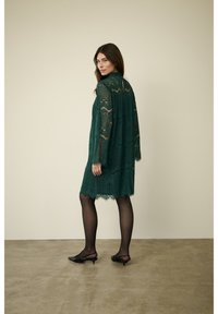 Zelené krajkové šaty s volným střihem, dlouhými rukávy, složitými vzory, s vykrojeným lemem, doplněné o černé boty na nízkém podpatku a neprůhledné punčochy.