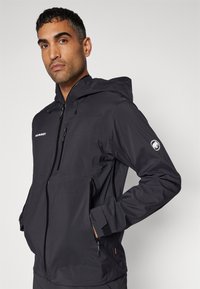 Schwarze technische Jacke mit Kapuze, vorderen Reißverschlüssen und Seitentaschen. Verfügt über elastische Bündchen und ein Logo-Patch an den Ärmeln.