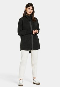 Schwarze Fleece-Jacke mit Reißverschluss und hohem Kragen, langen Ärmeln und kontrastierendem grauen Reißverschluss. Kombiniert mit weißen Hosen und lässigen Sneakers.