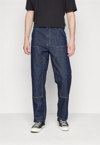 Dark blue denim wide-leg pants with contrasting yellow stitching, featuring large side pockets and a relaxed fit. Paired with black sneakers.