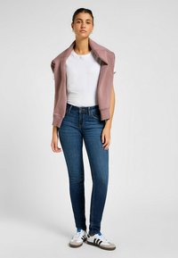 Jeans skinny blu, t-shirt bianca e una felpa rosa chiaro appoggiata sulle spalle. Le scarpe da ginnastica hanno una base bianca con strisce scure.
