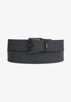 Zwarte gestructureerde leren riem met zwarte metalen gesp en "kazar" logo op de riemlus, plat opgerold tegen een witte achtergrond.