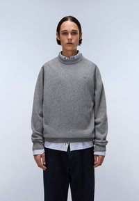 Maglione grigio a maglia con collo alto, vestibilità rilassata e polsini a costine, indossato sopra una camicia a righe blu e bianca. Pantaloni scuri visibili.