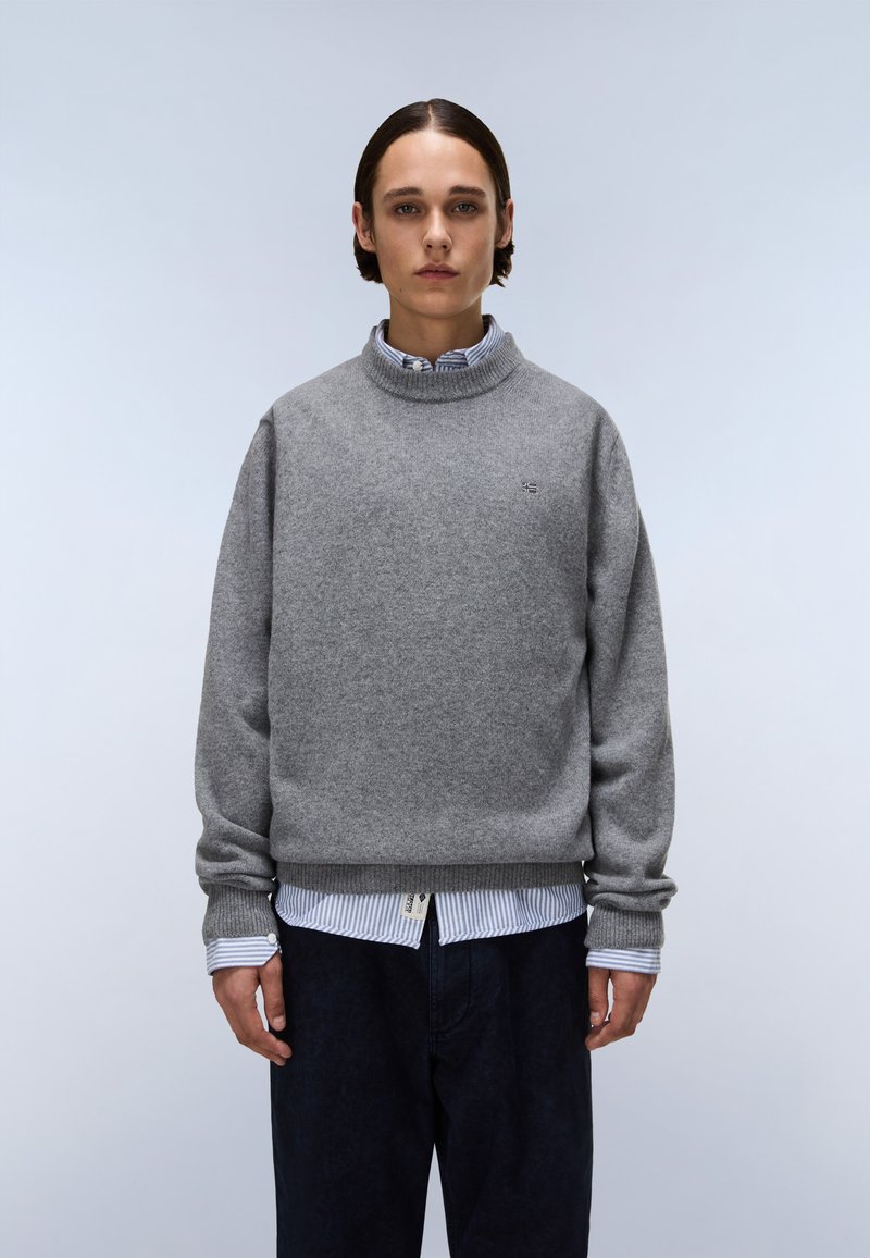Maglione grigio a maglia con collo alto, vestibilità rilassata e polsini a costine, indossato sopra una camicia a righe blu e bianca. Pantaloni scuri visibili.