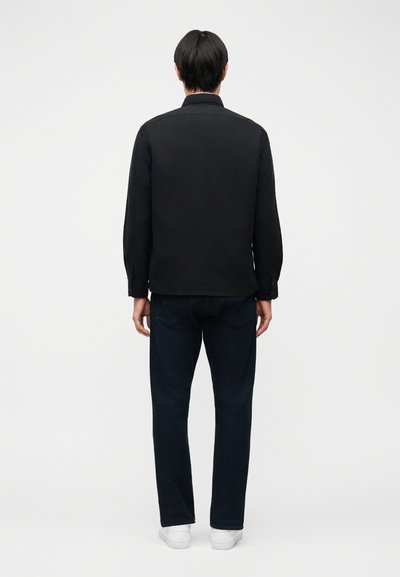 Man met zwart haar, gekleed in een zwart shirt met lange mouwen, donkere jeans en witte sneakers, staand met zijn rug naar de witte achtergrond gericht.