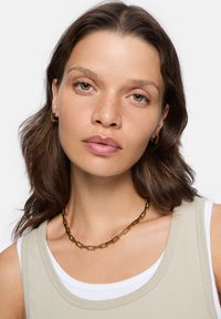 Guldkæde med rektangulære led, båret over en beige ribbet tanktop og hvid skjorte; modellen har let fregnet hud.