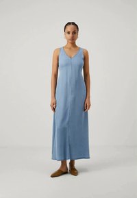 Femeie stând dreaptă, îmbrăcată într-o rochie maxi fără mâneci de culoare albastră deschisă și purtând pantofi plat de tip pointed de culoare maro, pe un fundal gri simplu.
