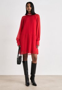 Crvena plisirana haljina s dugim prozirnim rukavima i visokim ovratnikom. Uparena s crnim čizmama do koljena s potpeticama i malom crnom torbicom.