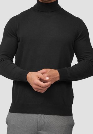 Homme portant un pull noir à col roulé ajusté et un pantalon gris, mains légèrement jointes à la taille devant un fond uni.