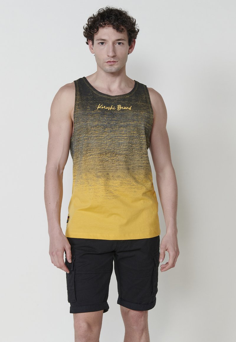 Črna in rumena gradientna majica brez rokavov z okroglim ovratnikom in logotipom "Karoshi Brand". Obuta z črnimi cargo hlacami z stranskimi žepi.