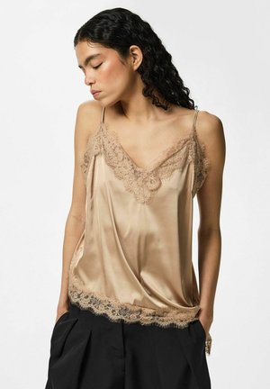 Kvinde med langt krøllet hår iført en beige satin camisole med blondekant og sorte højtaljede bukser, hænderne i lommerne, kigger ned.