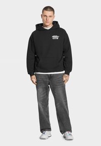 Czarna bluza z kapturem z przednią kieszenią i białym napisem graficznym, zestawiona z szarymi jeansami i białymi trampkami.