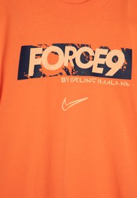 Oranžs kokvilnas t-kreklis ar lielu zilu un baltu grafiku, kas lasa "FORCE9" ar traipveida efektu un Nike logo apakšā.