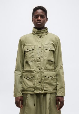 Személy világos olívazöld dzsekit visel, amelyen négy elöl zsebfedős zseb és magas gallér található, és egy egyszínű világos háttér előtt áll.