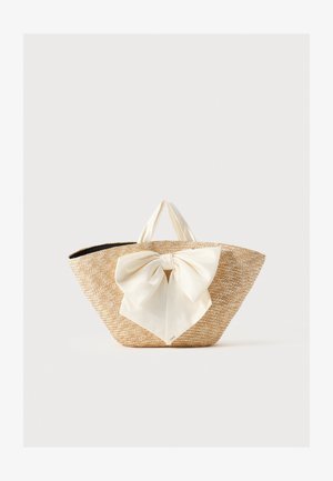 Sac cabas en paille tissée avec des poignées en tissu crème et un grand nœud décoratif crème à l'avant, sur un fond uni.