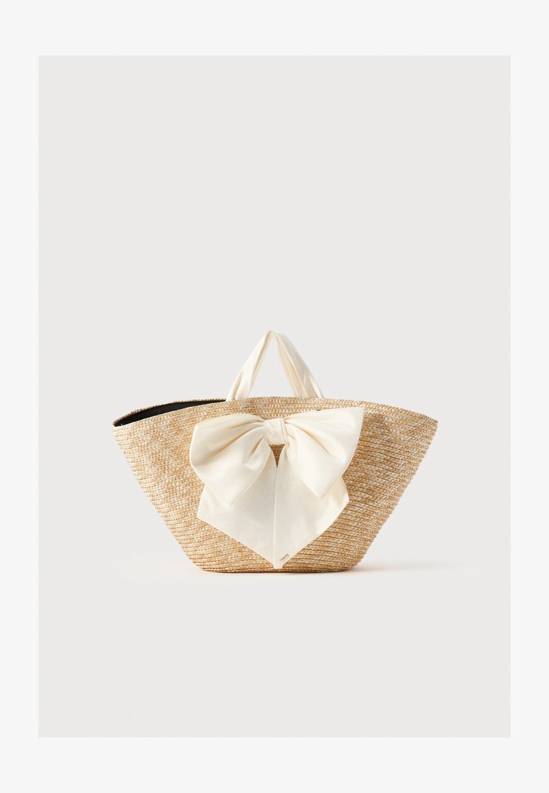 Sac cabas en paille tissée avec des poignées en tissu crème et un grand nœud décoratif crème à l'avant, sur un fond uni.