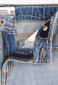 Jeans din denim albastru deschis cu fermoar parțial desfăcut, închise cu un nasture și cu eticheta vizibilă care citește "jeanși largi pentru copii zombie".