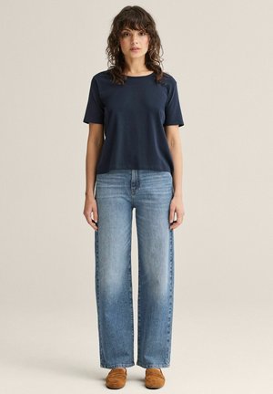 Kvinde med krøllet hår iført navyblå kortærmet skjorte, lyseblå bukser med brede ben og brune loafers, stående mod en beige baggrund.