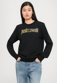 Donna con lunghi capelli scuri che indossa una felpa nera con il testo dorato "Just Cavalli" e jeans azzurri chiari, con le mani in tasca, su uno sfondo neutro.