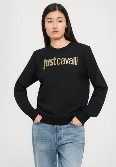 Femme avec de longs cheveux noirs portant un sweatshirt noir avec le texte doré "Just Cavalli" et un jean bleu clair, les mains dans les poches, devant un fond uni.