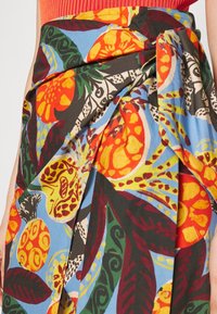 Jupe portefeuille à imprimé tropical lumineux avec de gros fruits orange, jaune et verts ainsi que des feuilles sur un fond bleu clair, portée avec un haut rouge.