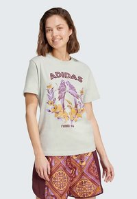 Šviesiai žalia medvilninė marškinėliai su grafika, kurioje pavaizduoti du paukščiai, apsupti geltonų gėlių, ir užrašu "ADIDAS FARM RA". Trumpi rankovės.