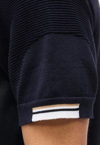 Κοντινό πλάνο μπλε ναυτικού πλεκτού πουκάμισου με κοντό μανίκι, με ριμπ υφή στον ώμο και λευκές, μαύρες και μπεζ ρίγες στη μανσέτα του μανικιού.