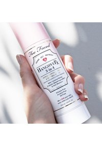 Too Faced Hangover 3-in-1 spray in een roze cilindervormige fles, met witte accenten en een label met productdetails in zwarte en gouden tekst.