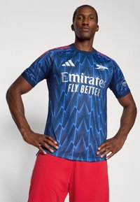 Blå fodboldtrøje med lynmønster, der har hvid sponsorlogo "Emirates" og tre røde striber på skuldrene. Korte ærmer, rund hals.