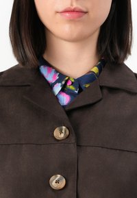 Žena s tamnosmeškom bob frizurom nosi smešani smešani smešan jaknu s dugmadima preko živopisne košulje s cvjetnim uzorkom.