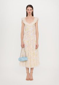 Vrouwelijke gebloemde jurk met pastelkleuren, transparante stof, ruches bij de hals, gecombineerd met een lichtblauwe handtas en beige hakken met banden.