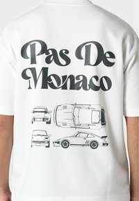 Camiseta blanca con "Pas De Monaco" en negrita de color negro y ilustraciones gráficas de varios coches en diferentes orientaciones.