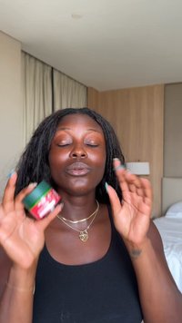 Bocal rouge et vert avec un couvercle noir tenu dans les mains ; texture lisse ; étiquettes visibles ; personne portant un débardeur noir avec des colliers superposés.