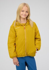 Veste isolante jaune avec une capuche, fermeture éclair avant beige, poignets ajustables et logo sur le côté gauche de la poitrine. Associée à un pantalon bleu.