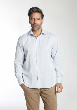 Homme portant une chemise à manches longues à carreaux bleu clair et un pantalon beige, debout les mains dans les poches sur un fond uni.