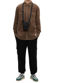 Person in braunem Button-Up-Hemd, schwarzen Cargo-Hosen, türkis-weißen Sneakers und einer kleinen schwarzen Umhängetasche mit Händen in den Taschen.