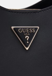 Il materiale nero testurizzato presenta un emblema triangolare dorato e nero ben visibile con il logo "GUESS" e un punto interrogativo.