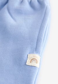 Lys blå strikket stof med et foldet design, der har en tekstureret lap med et regnbue-motiv i beige og lys brune farver.