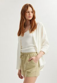 Camisa blanca de botones sobre una camiseta sin mangas blanca de canalé, combinada con unos pantalones cortos beige claros con cordón y bolsillos laterales. Textura suave.