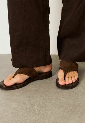 Füße in dunkelbraunen Wildledersandalen mit breiten Riemen, kombiniert mit lockeren braunen Hosen, stehend auf einem grauen Betonboden.