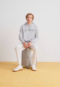 Lichtgrijze hoodie met zwart logo, casual witte broek en witte sneakers met lichtbruine zolen, zittend op een zilveren koffer.