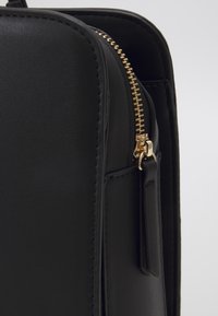 Sac en similicuir noir à texture lisse, muni d'une fermeture éclair dorée et d'une languette, forme rectangulaire et design minimaliste.