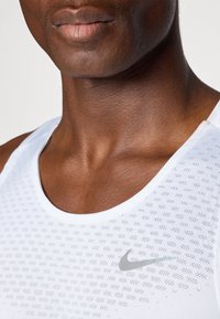 Valkoinen Nike-sifonkipaita, jossa on pyöreä kaula-aukko, litteät saumat ja tekstuuroitu kangas, jossa on pieniä reikiä. Harmaa Nike-logo rinnassa.