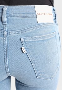 Vaqueros de mezclilla en un tono azul claro con un diseño ajustado. Las características incluyen una etiqueta de cuero blanco en la cinturilla y una pequeña etiqueta de marca en el bolsillo trasero.